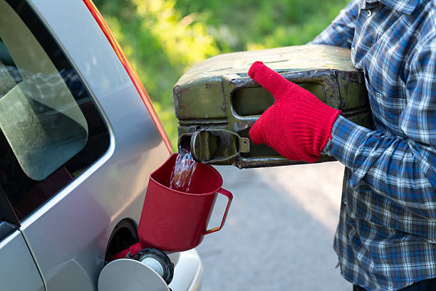 A man in red gloves pours gasoline into a gray car fuel tank from a canister using a funnel